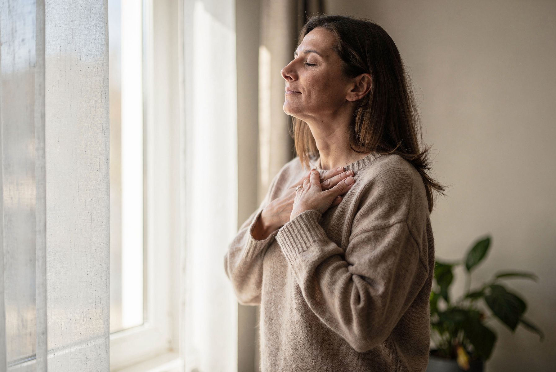 Eine Frau steht am Fenster, die Hände auf der Brust, und atmet ruhig aus.