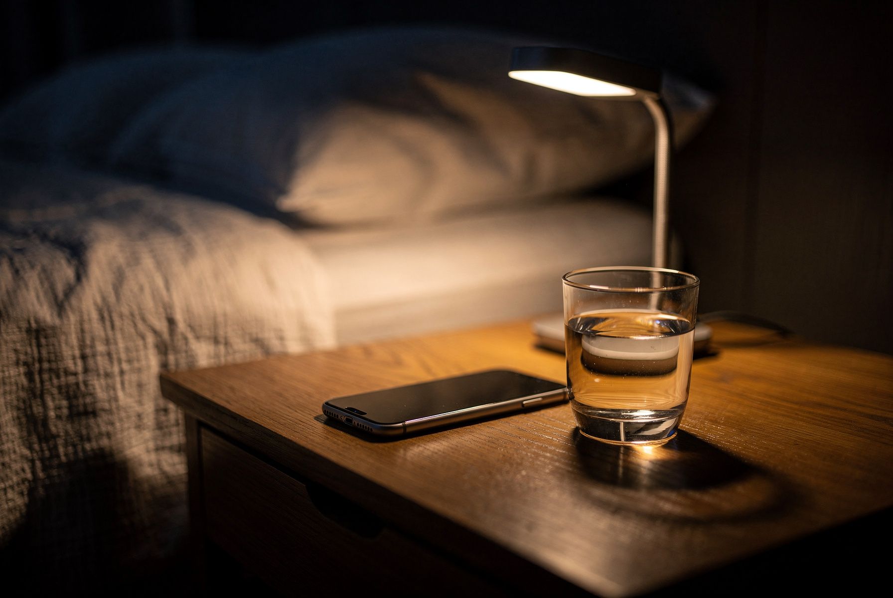 Nachttisch am Abend mit umgedrehtem Handy, Leselampe und einem Glas Wasser.