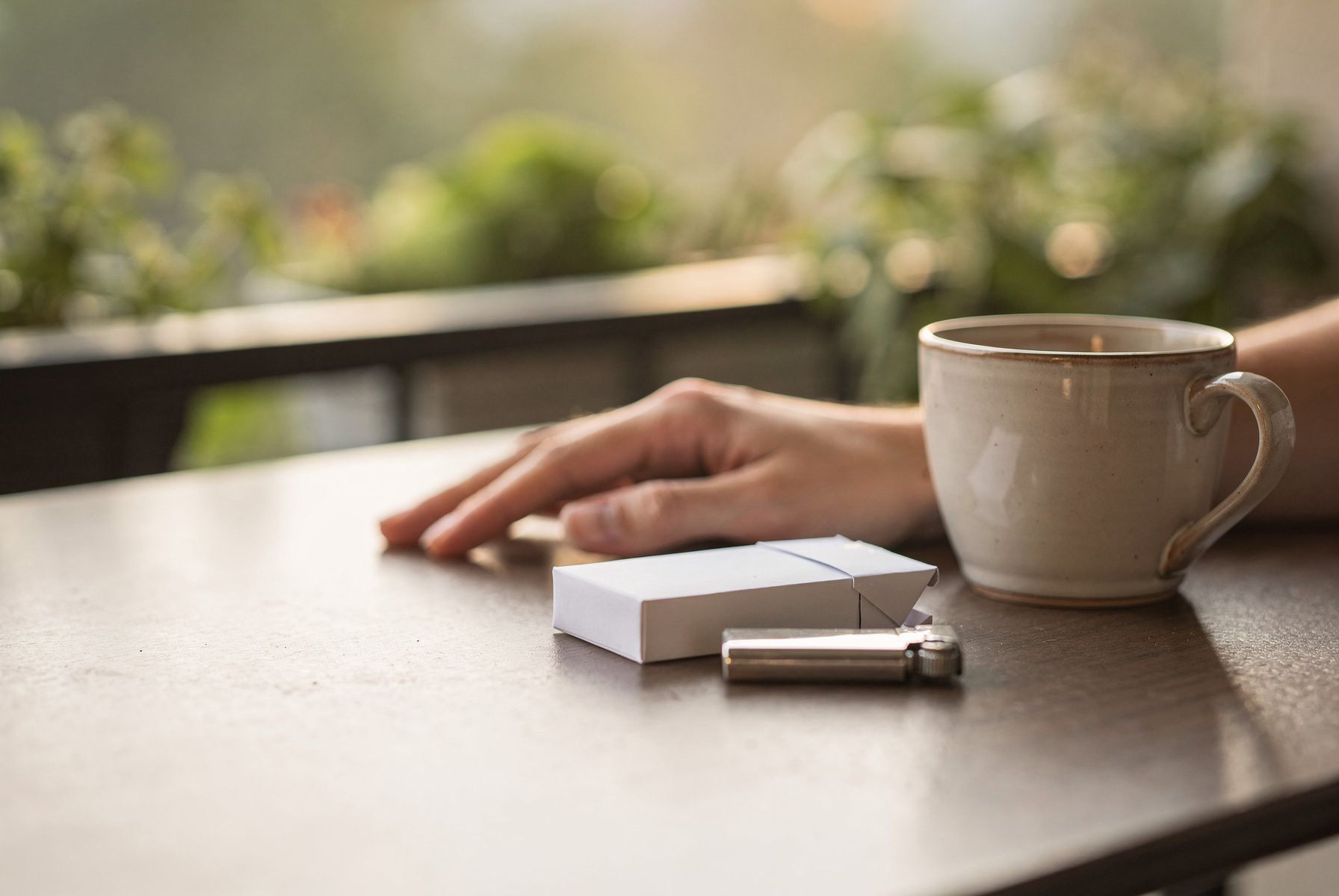 Balkontisch am Morgen mit Zigarettenschachtel, Feuerzeug und Kaffee; eine Hand greift nicht zu.