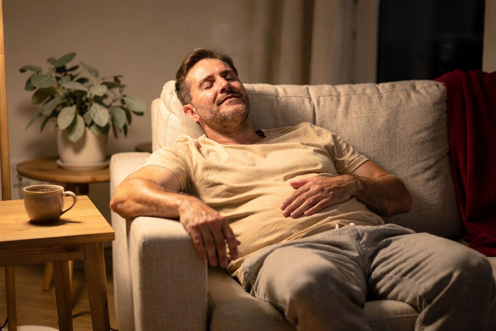 Ein Mann liegt am Abend entspannt auf dem Sofa, eine Hand auf dem Bauch, daneben eine Tasse Tee.
