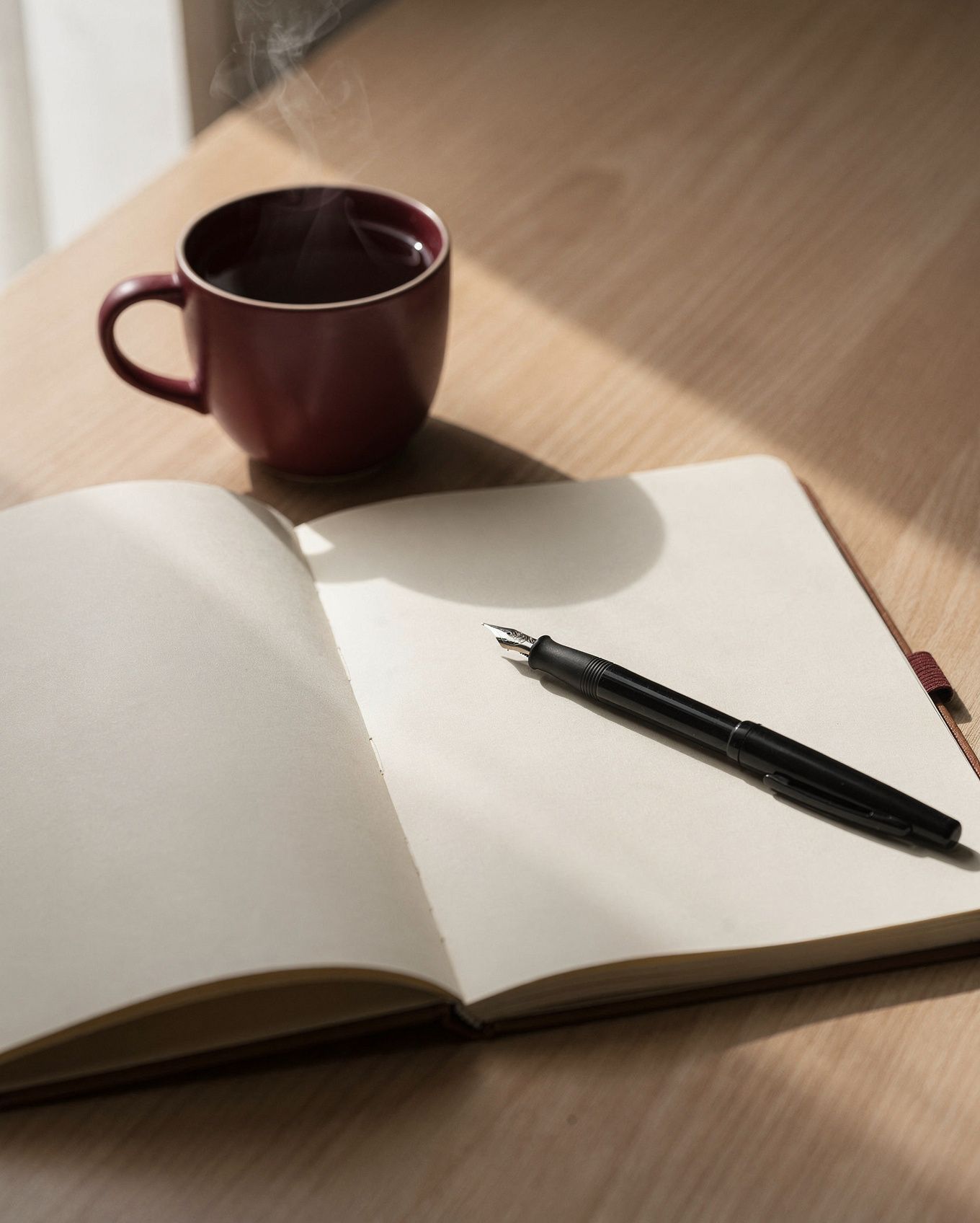 Ein offenes Notizbuch, ein Füller und eine weinrote Tasse auf einem Holztisch.