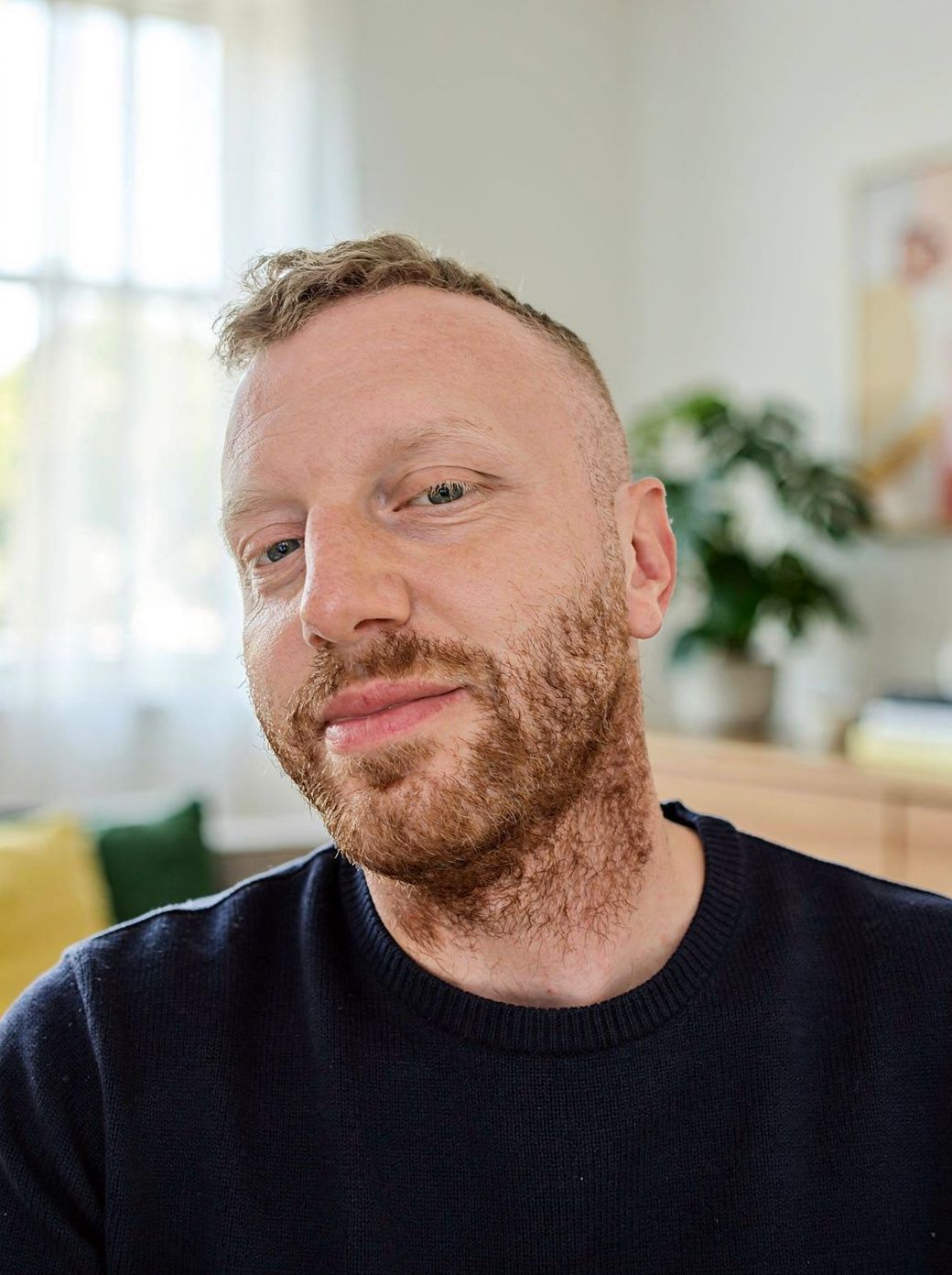 Tommy Müller im dunkelblauen Pullover, lächelnd, in einem hellen Wohnzimmer.